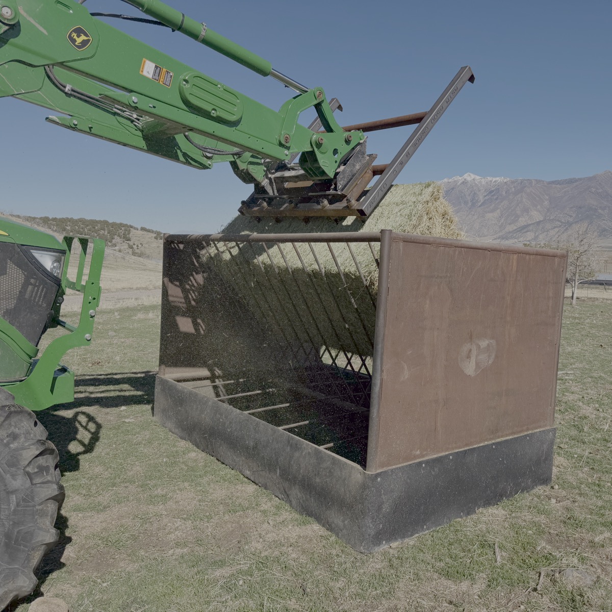 Green excavator lifting hay into a metal container with mountains in the background